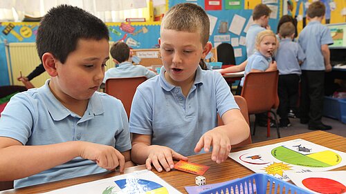 Two children sitting at a desk playing a game, another looks over from afar.